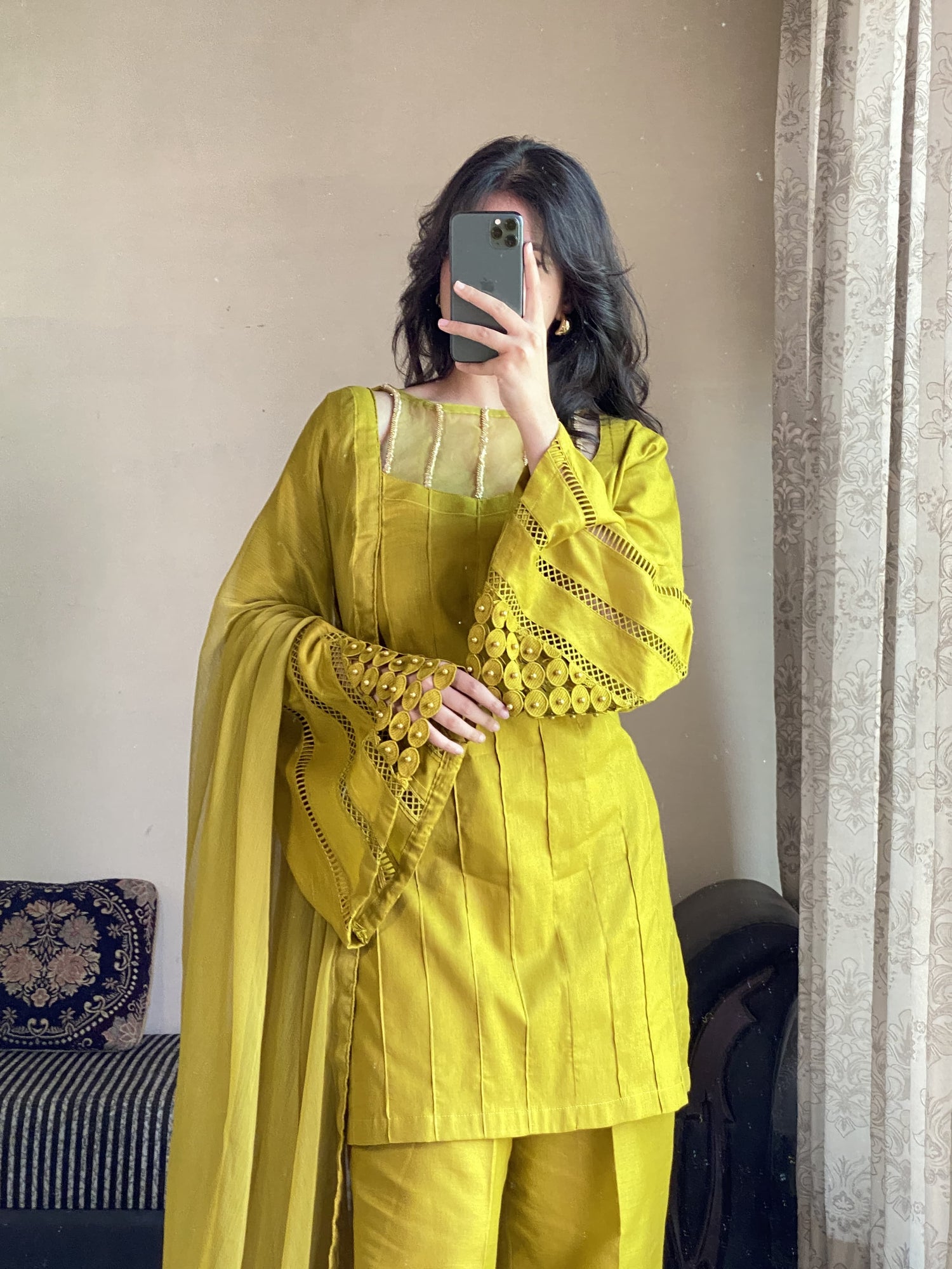 Female wearing a yellow traditional outfit with intricate designs, standing in front of a neutral-colored wall.